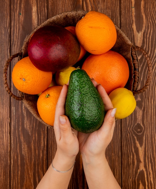 木製のテーブル上のバスケットにアボカドマンゴーレモンオレンジとしてアボカドと柑橘系の果物を保持している女性の手の上から見る