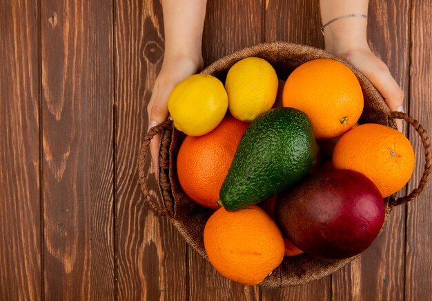 Взгляд сверху рук женщины держа корзину полный цитрусовых фруктов как апельсин лимона манго авокадоа на деревянном столе с космосом экземпляра