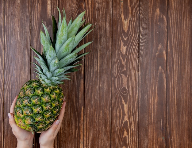 Top view of woman hands holding pineapple on left side and wooden background with copy space