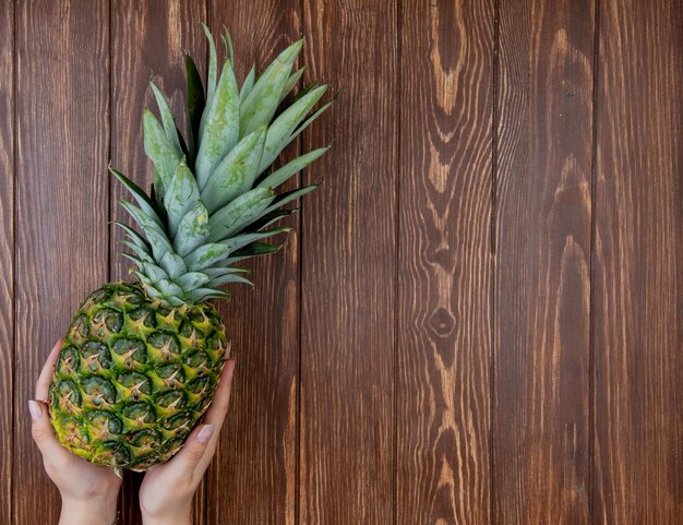 Top view of woman hands holding pineapple on left side and wooden background with copy space