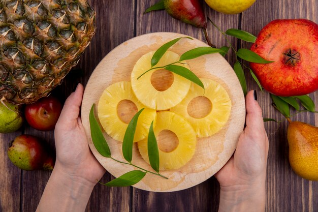 Top view of woman hands holding pineapple slices on cutting board with pineapple peach plum pomegranate on wooden surface