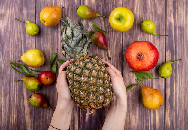 Top view of woman hands holding pineapple with apple peach plum pomegranate on wooden surface