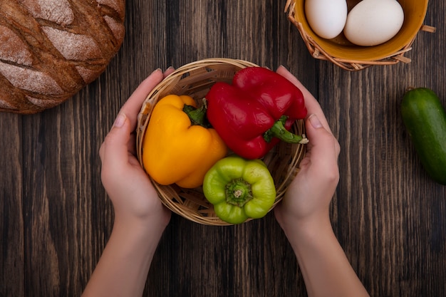 Foto gratuita donna di vista superiore che tiene peperoni colorati in un cestino con uova di gallina e pane nero su fondo di legno