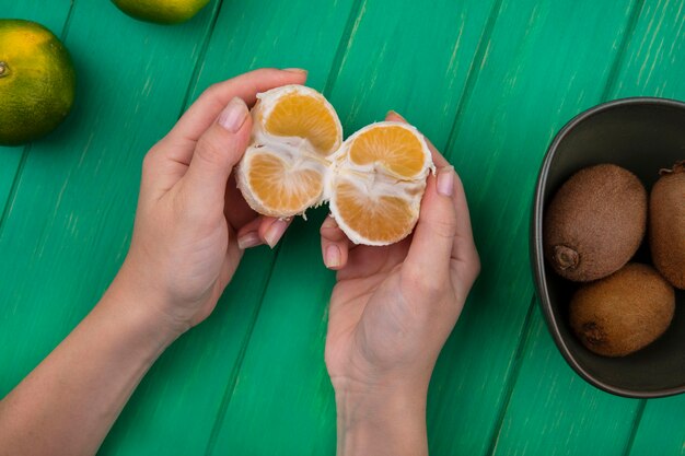 Top view woman holding peeled tangerine in her hands with kiwi in a bowl on a green wall