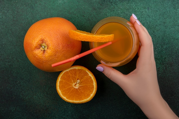 Top view  a woman holds in her hand orange juice in a glass with a red straw and a slice of orange with grapefruit on a green background