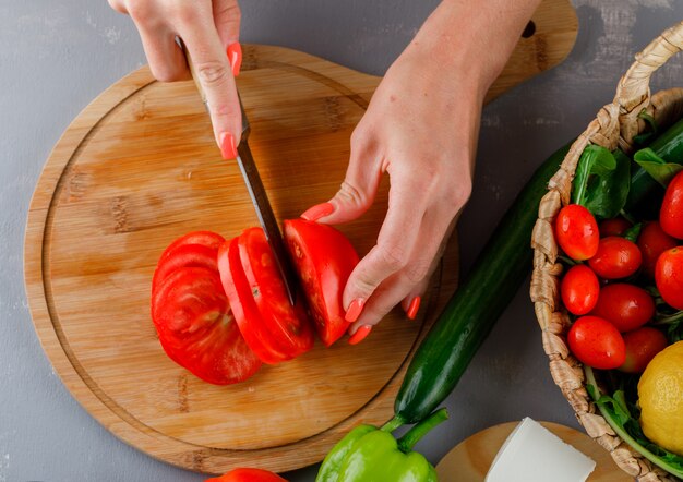 Top view woman slicing tomato on cutting board with cucumber, green pepper on gray surface