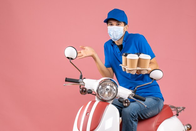 Top view of wondering courier man in mask wearing hat sitting on scooter showing orders on pastel peach