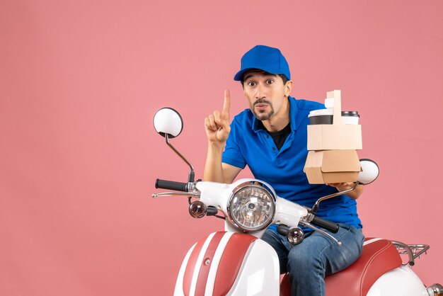 Top view of wondering courier man wearing hat sitting on scooter showing orders pointing up on pastel peach