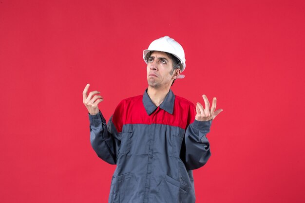 Top view of wondering young builder in uniform wearing hard hat on isolated red wall