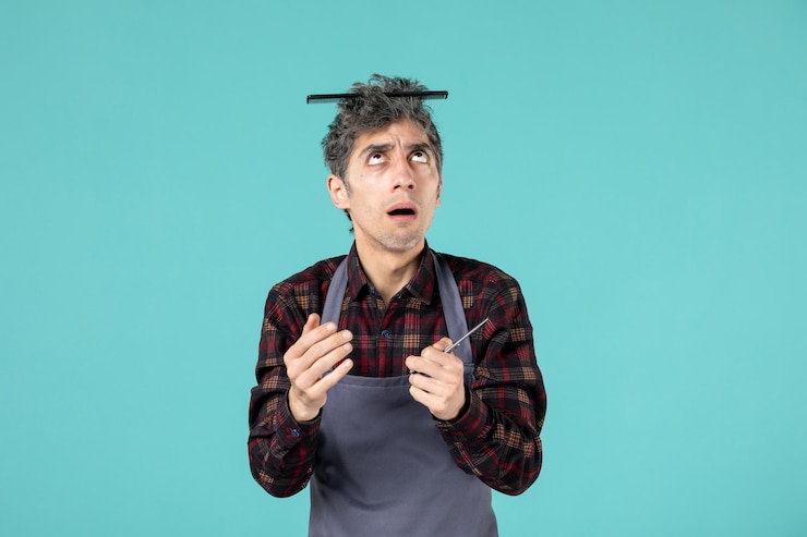 Free Photo top view of wondering young hairdesser wearing gray apron and holding comb on his head and showing scissor on pastel blue color background