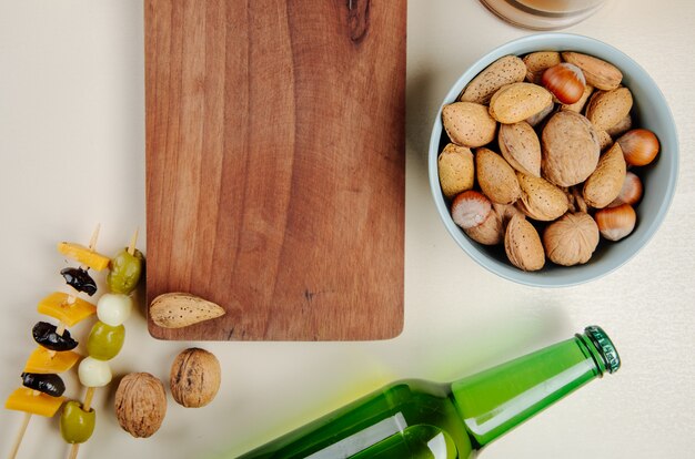 Top view of a wood board and mix of nuts in a bowl pickled olives and bottle of beer on white