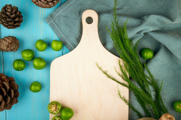 Top view of a wooden board and scattered sour green plums with cones on blue wooden table