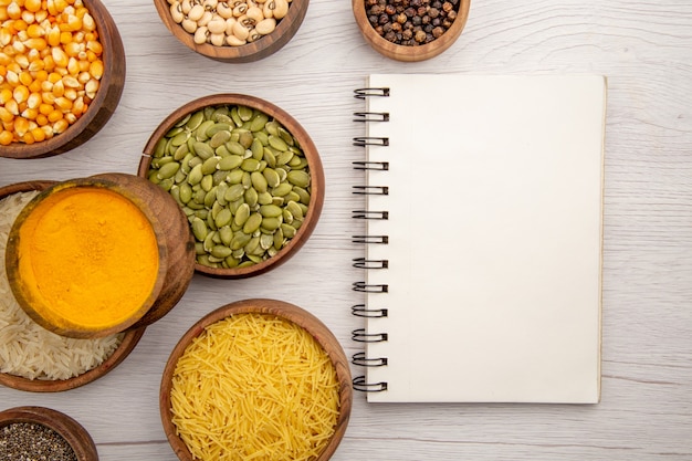 Top view wooden bowls with rice black pepper beans pumpkin seeds turmeric thin pasta tubes notebook on grey table