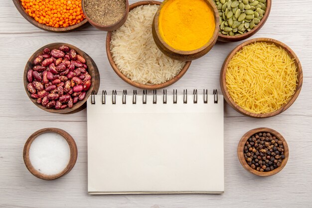 Top view wooden bowls with rice black pepper beans pumpkin seeds turmeric thin pasta tubes salt lentiles notepad on grey table