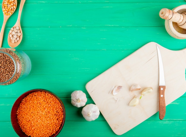 Top view of wooden cutting board with knife and garlic and raw red lentils in a bowl on green background with copy space