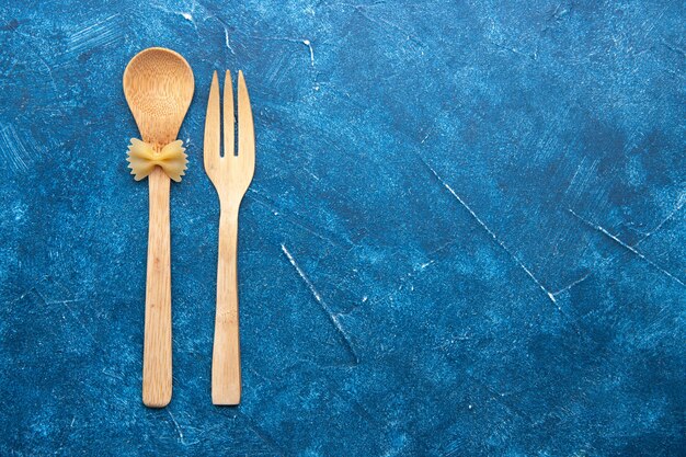 Top view wooden fork spoon farfalle on spoon on blue table with free space