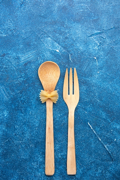 Top view wooden fork spoon farfalle on spoon on blue table with free space