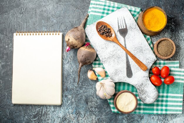 Top view wooden spoon on cutting board spices in bowls cherry tomatoes garlic beets notebook on grey table