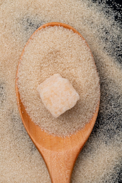 Top view of a wooden spoon with brown sugar and sugar cube on granulated sugar background