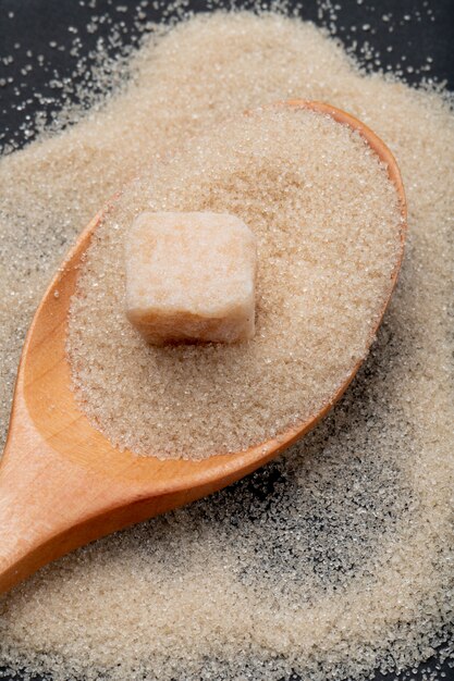 Top view of a wooden spoon with granulated brown sugar and sugar cube on black background