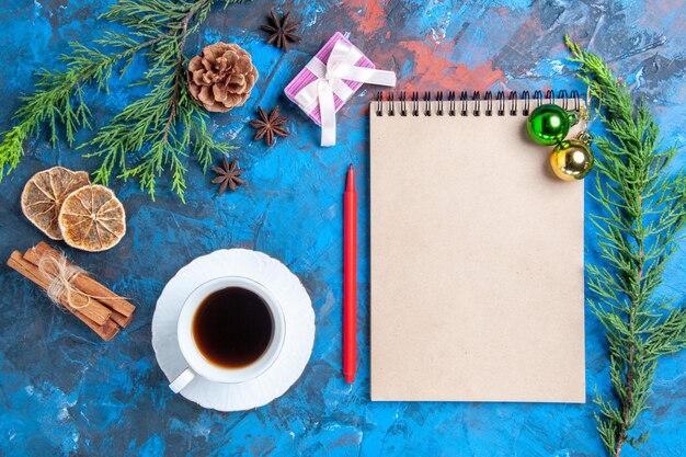 Top view xmas balls on a notebook pine tree branches cinnamon sticks anises dried lemon slices a cup of tea on blue surface