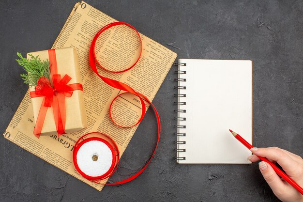 Top view xmas gift in brown paper branch fir ribbon on newspaper notepad pencil in female hand on dark surface