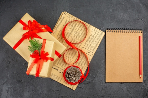 Top view xmas gift in brown paper branch fir ribbon on newspaper pinecone a notepad pencil on dark surface