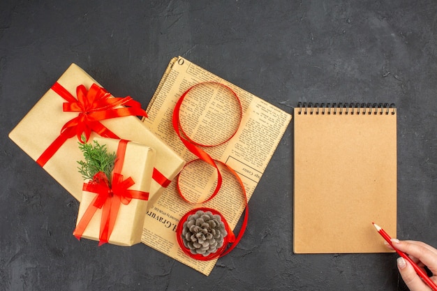 Top view xmas gift in brown paper branch fir ribbon on newspaper pinecone notepad red pencil in female hand on dark surface