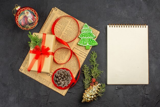 Top view xmas gift in brown paper branch fir ribbon on newspaper xmas ornaments a notebook on dark surface