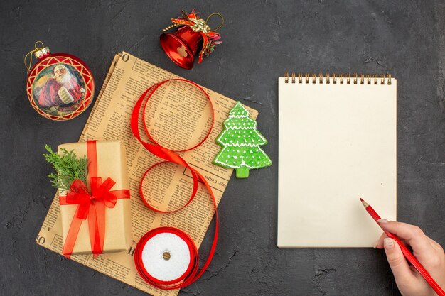 Top view xmas gift in brown paper branch fir ribbon on newspaper xmas ornaments notepad pencil in female hand on dark surface