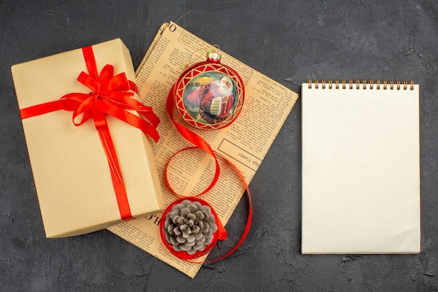 Top view xmas gift in brown paper ribbon xmas tree toy on newspaper a notebook on dark surface