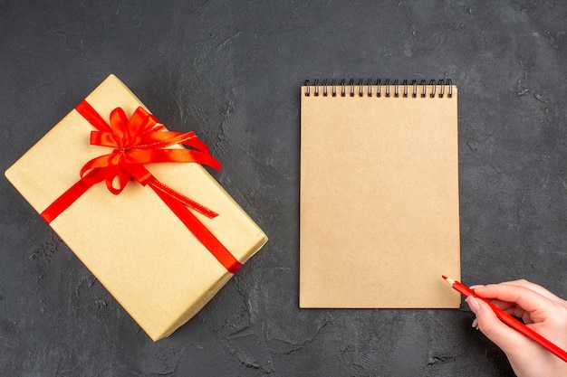 Top view xmas gift in brown paper tied with red ribbon notebook pen in female hand on dark surface