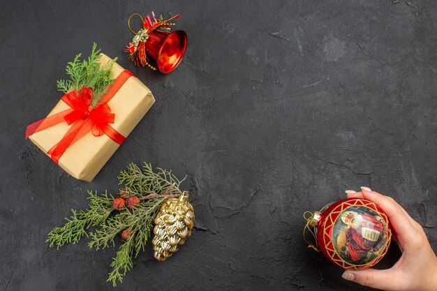 Top view xmas gift in brown paper tied with red ribbon xmas tree ornaments xmas ball in female hand on dark surface