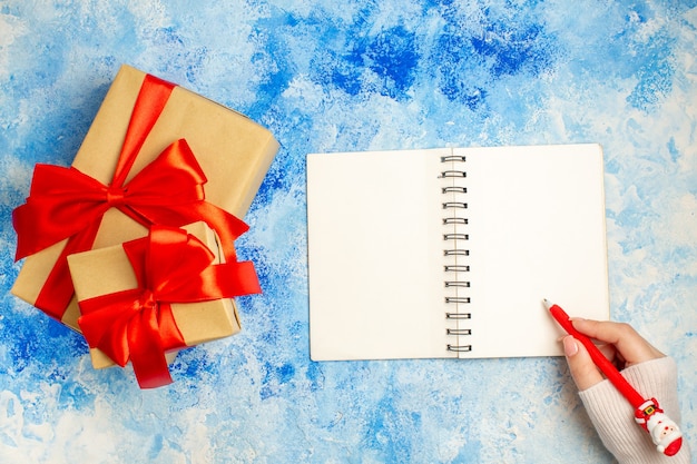Вид сверху рождественский подарок, перевязанный красным бантом, блокнот, ручка санта-клауса в женской руке на синем столе