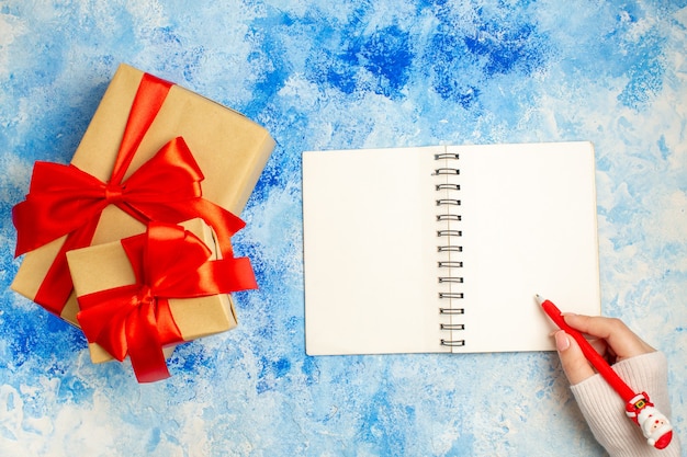 Top view xmas gift tied with red bow notepad santa pen in female hand on blue table