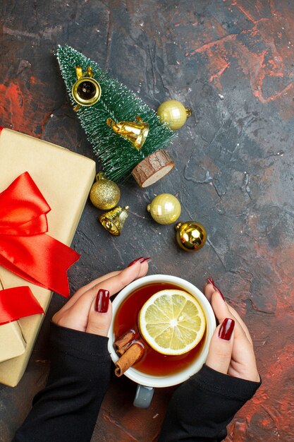 Top view xmas gifts golden xmas balls cup of tea in woman hand mini xmas tree on dark red table