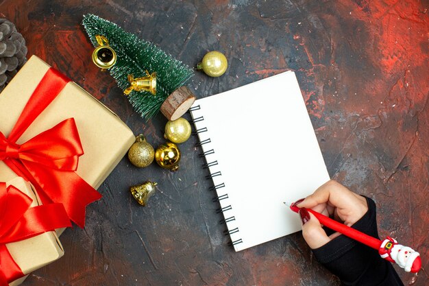 Top view xmas gifts golden xmas balls mini xmas tree notebook pen in woman hand on dark red table