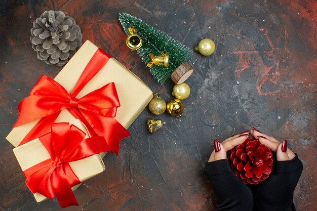 Top view xmas gifts golden xmas balls red pinecone in woman hands small xmas tree on dark red table