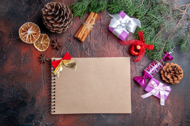 Top view xmas gifts pine tree branches with cone xmas tree toys cinnamon dried lemon slices anises a notebook on dark red surface