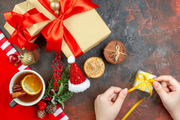 Вид сверху рождественские подарки, перевязанные красной лентой, шляпа санта-клауса, печенье, мини-подарок в женской руке, чашка чая на темно-красном столе