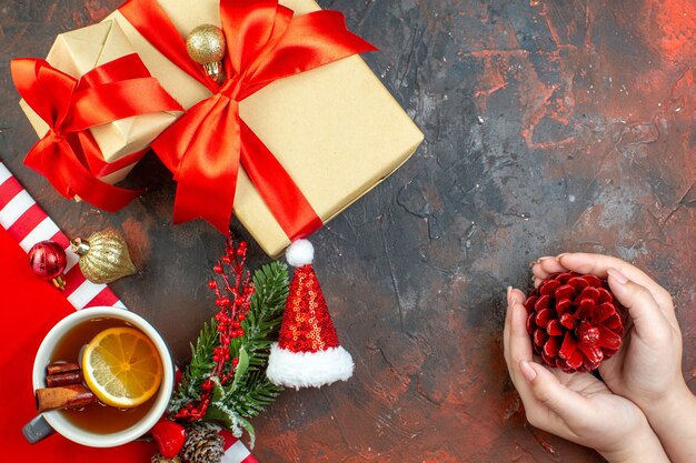 Top view xmas gifts tied with red ribbon santa hat pinecone in female hand cup of tea on dark red table