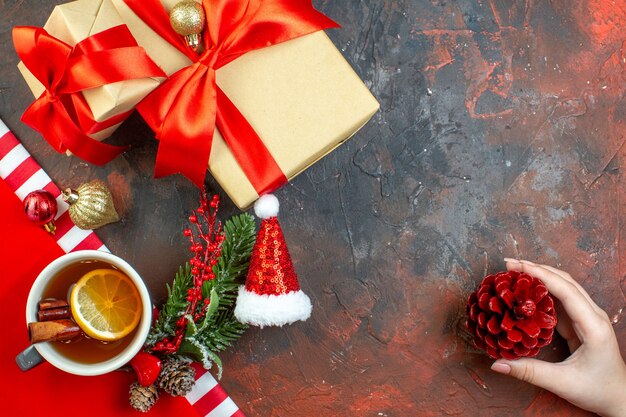 Top view xmas gifts tied with red ribbon santa hat pinecone in female hand on dark red table copy place