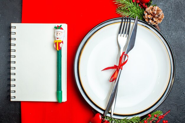 Вид сверху на рождественский фон с набором столовых приборов с красной лентой на аксессуарах для украшения обеденной тарелки еловые ветки рядом с блокнотом с ручкой на красной салфетке