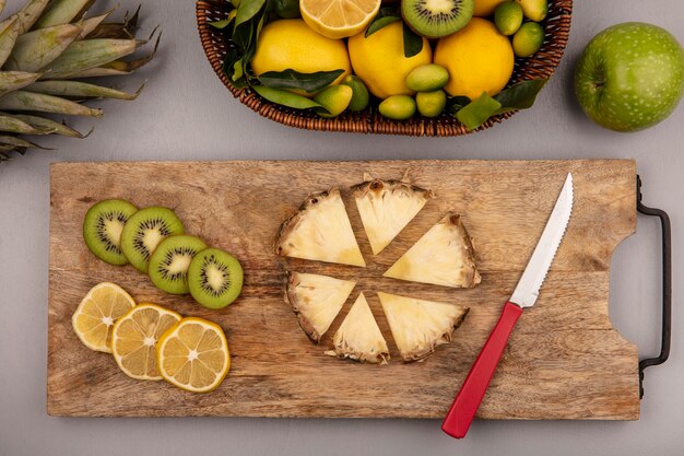 Top view of yellow and green fruits such as kiwi kinkans and lemons on a bucket with slices of kiwi lemon and pineapple on a wooden kitchen board with knife on a grey background