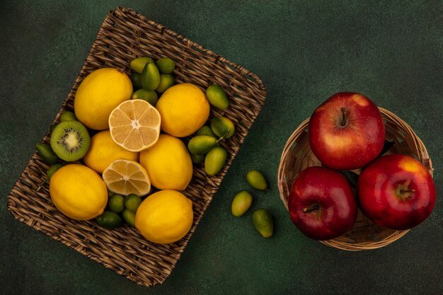 Top view of yellow lemons on a wicker tray with kinkans with red apples on a bucket on a green wall