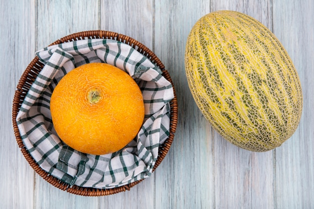 Top view yellow melon on a checked tablecloth on a bucket with whole cantaloupe on grey wooden surface