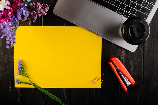 Top view of yellow paper sheet with colorful paper clips with purple color statice flowers and laptop with a cup of coffee red stapler on dark wooden background
