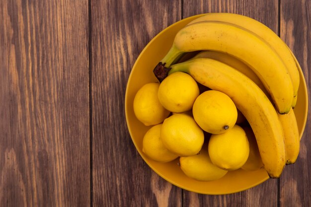 Top view of yellow skinned lemons on a yellow plate with bananas on a wooden surface with copy space