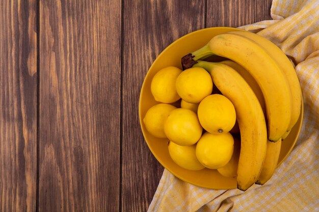 Top view of yellow skinned lemons on a yellow plate on a yellow checked cloth with bananas on a wooden surface with copy space