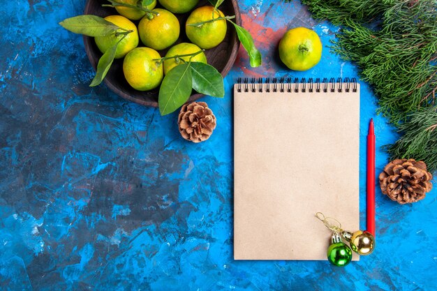 Top view yellow tangerines with leaves in wooden bowl a notebook and red pencil xmas ornaments on blue surface